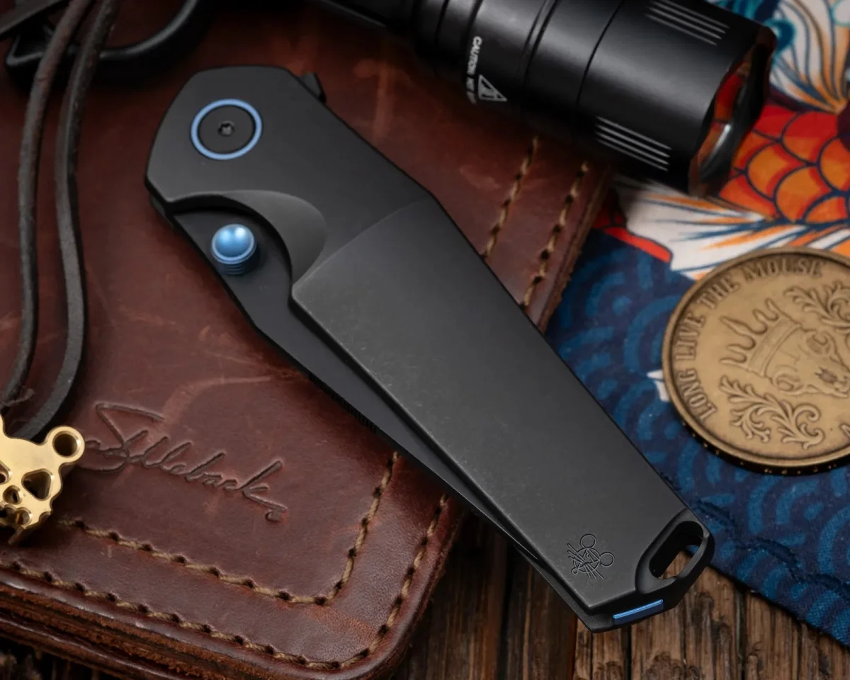 Black folding pocket knife featuring blue pivot and thumb stud accents, resting on a leather wallet alongside a flashlight and a metal coin.