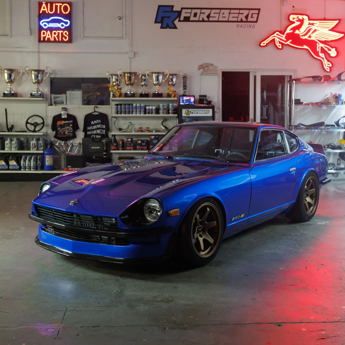 Blue Datsun 280Z with bronze wheels parked inside a garage decorated with neon signs and trophies.