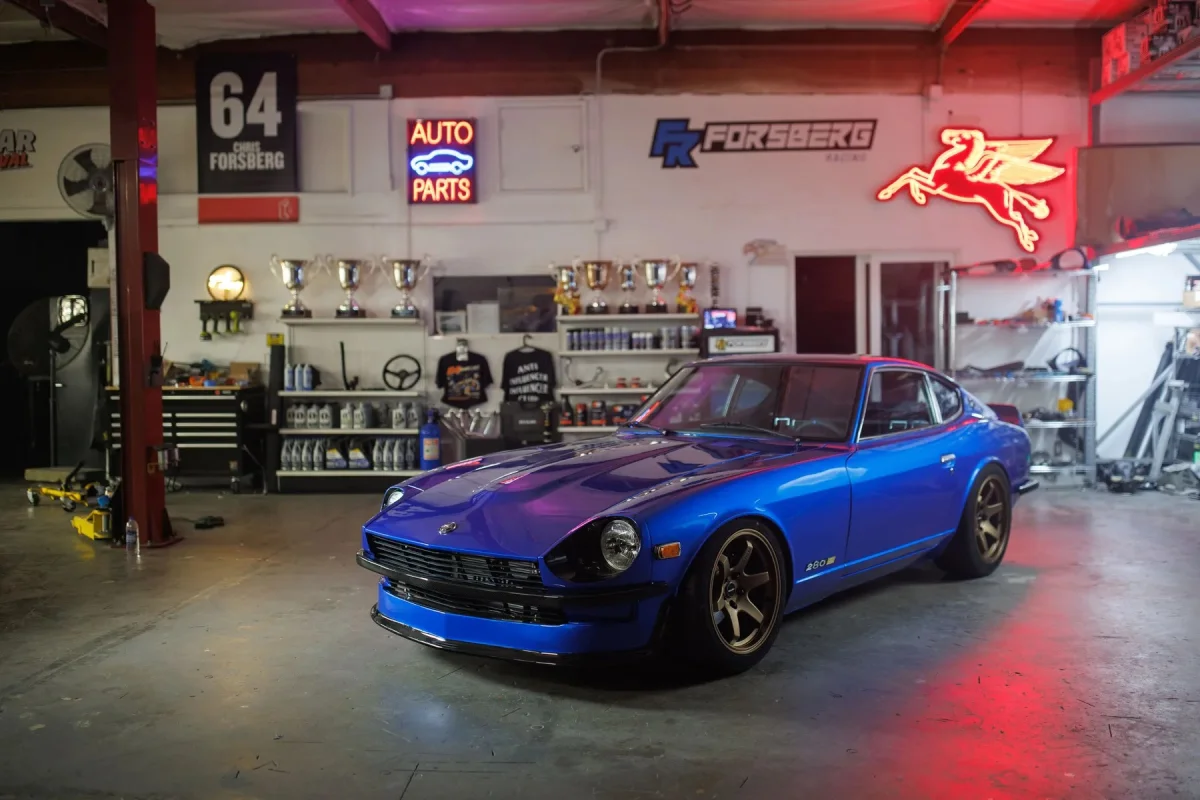 Blue Datsun 280Z with bronze wheels parked inside an automotive garage decorated with racing trophies and neon signs.