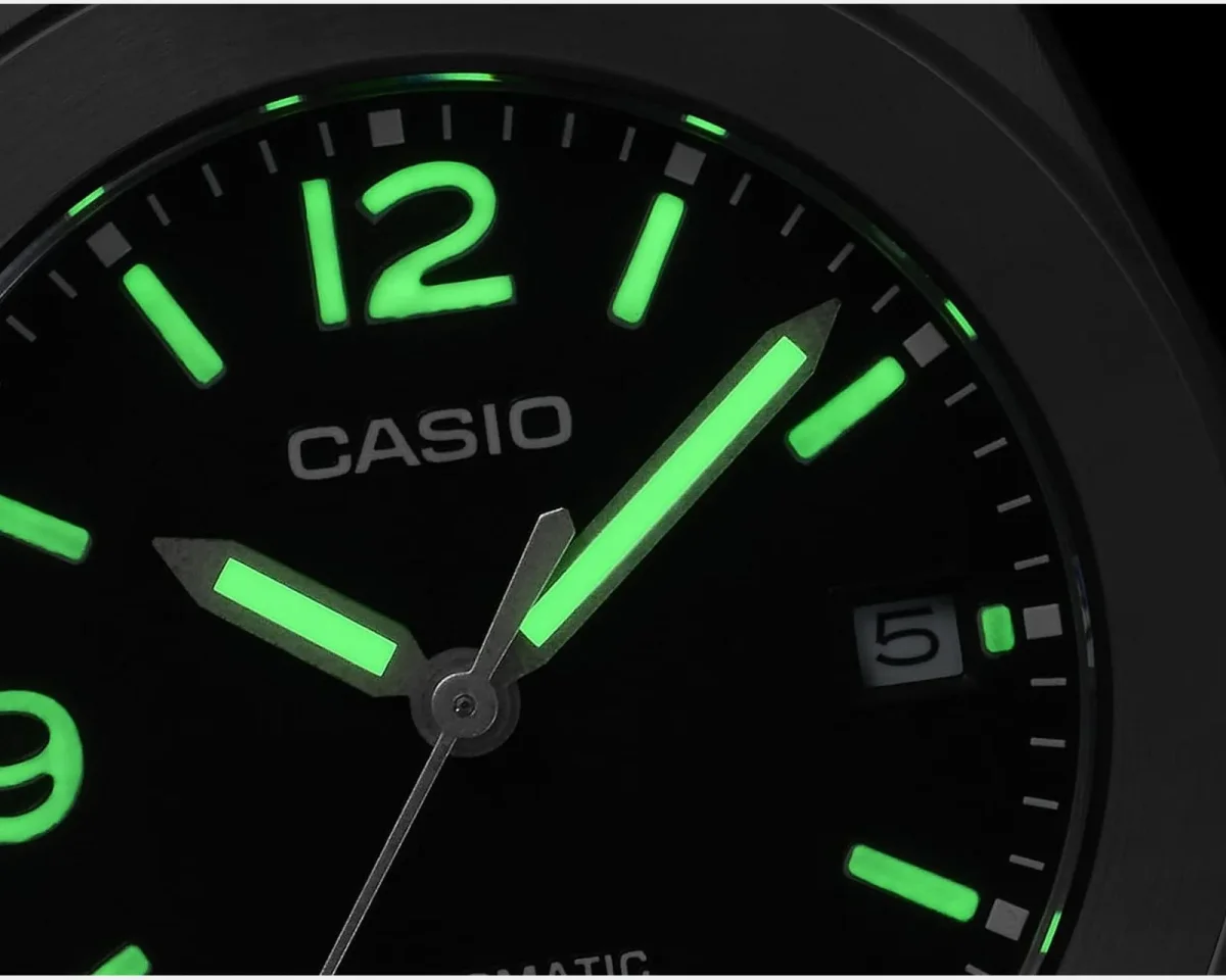 Close-up of a black Casio watch face with glowing green luminous hands and hour markers in the dark.