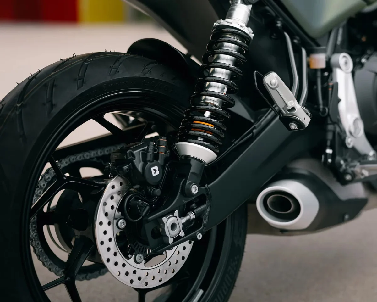 Close-up of a motorcycle rear wheel, featuring the suspension coil-over shock absorber, disc brake, chain drive, and exhaust pipe.