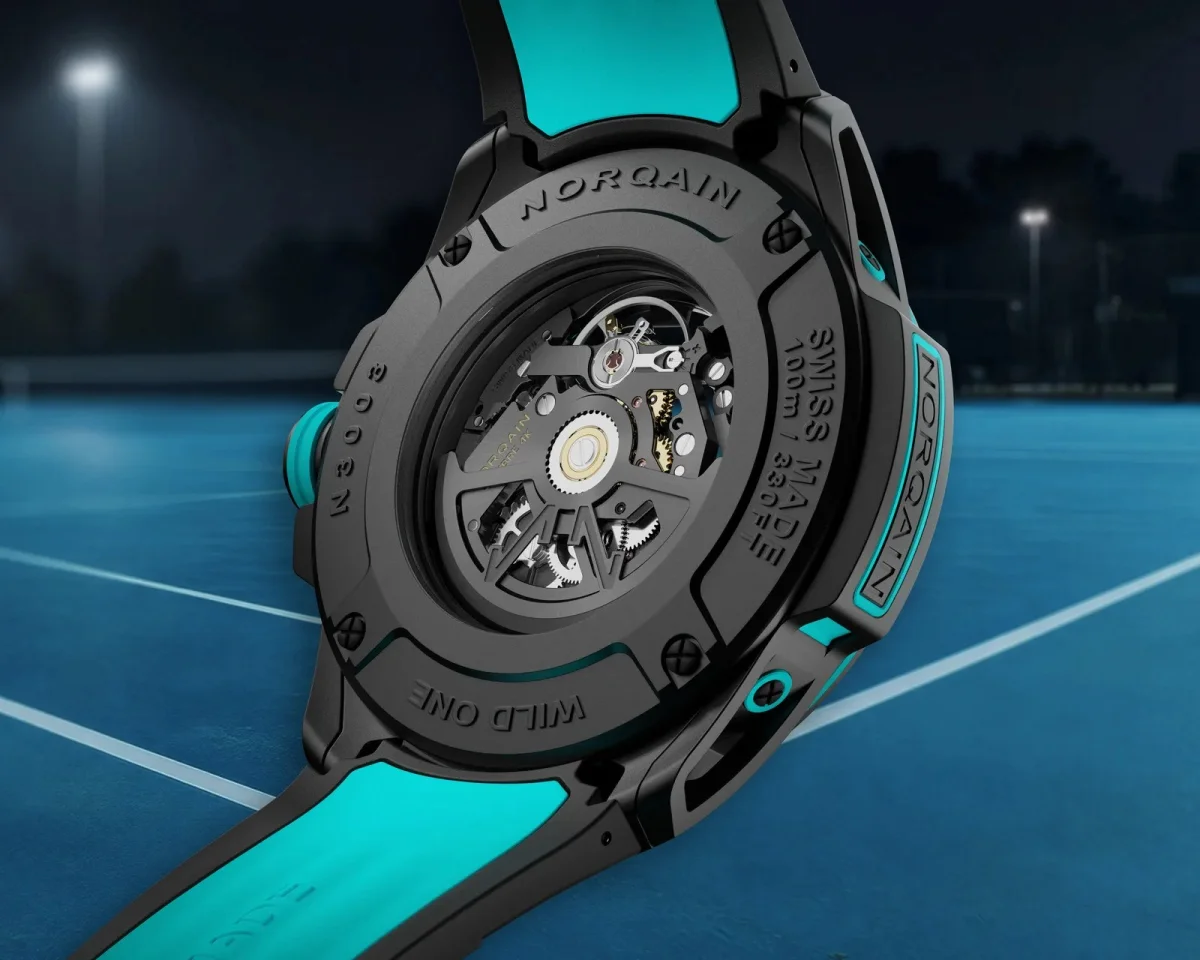 Back of a Norqain Wild One watch featuring a black case, turquoise accents, and an open caseback revealing the mechanical movement, positioned against a blue tennis court.
