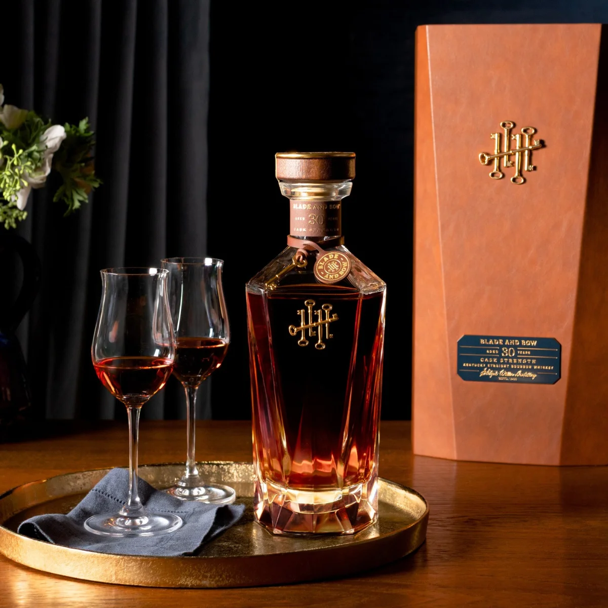 Blade and Bow 30-Year Kentucky Straight Bourbon Whiskey in a faceted glass decanter with two tasting glasses on a gold tray, positioned next to its leather-wrapped presentation box.