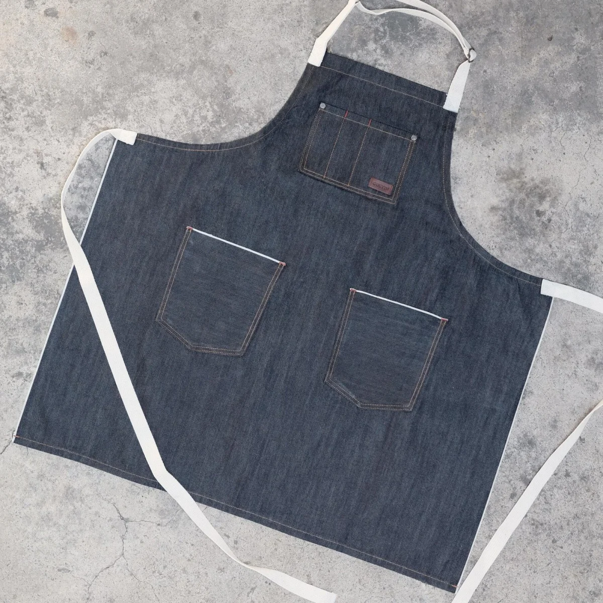 Dark blue denim apron with white straps, a chest pocket, and two waist pockets, displayed on a concrete surface.