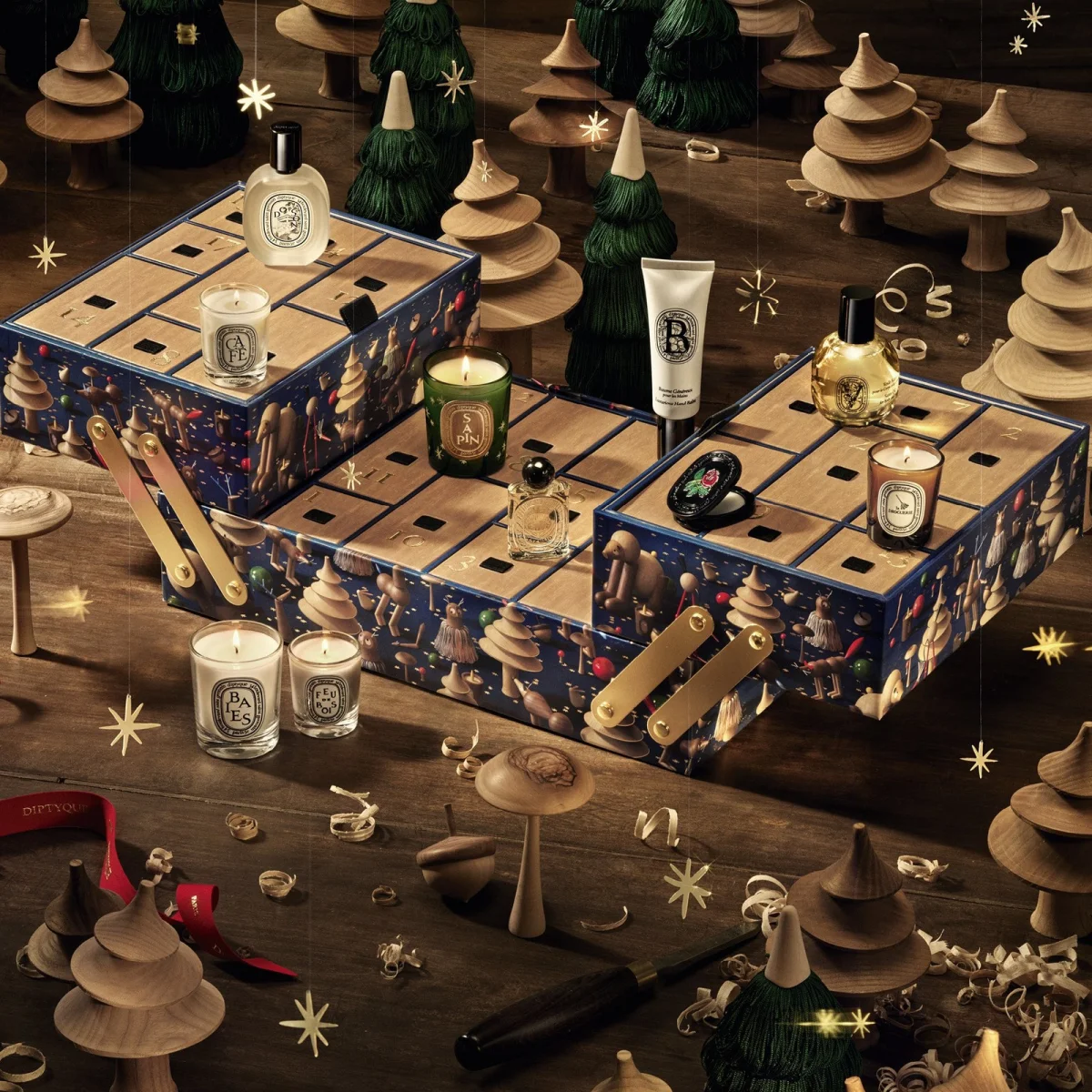 Diptyque advent calendar box filled with assorted candles, fragrances, and a hand cream, arranged on a wooden floor among decorative wooden trees.