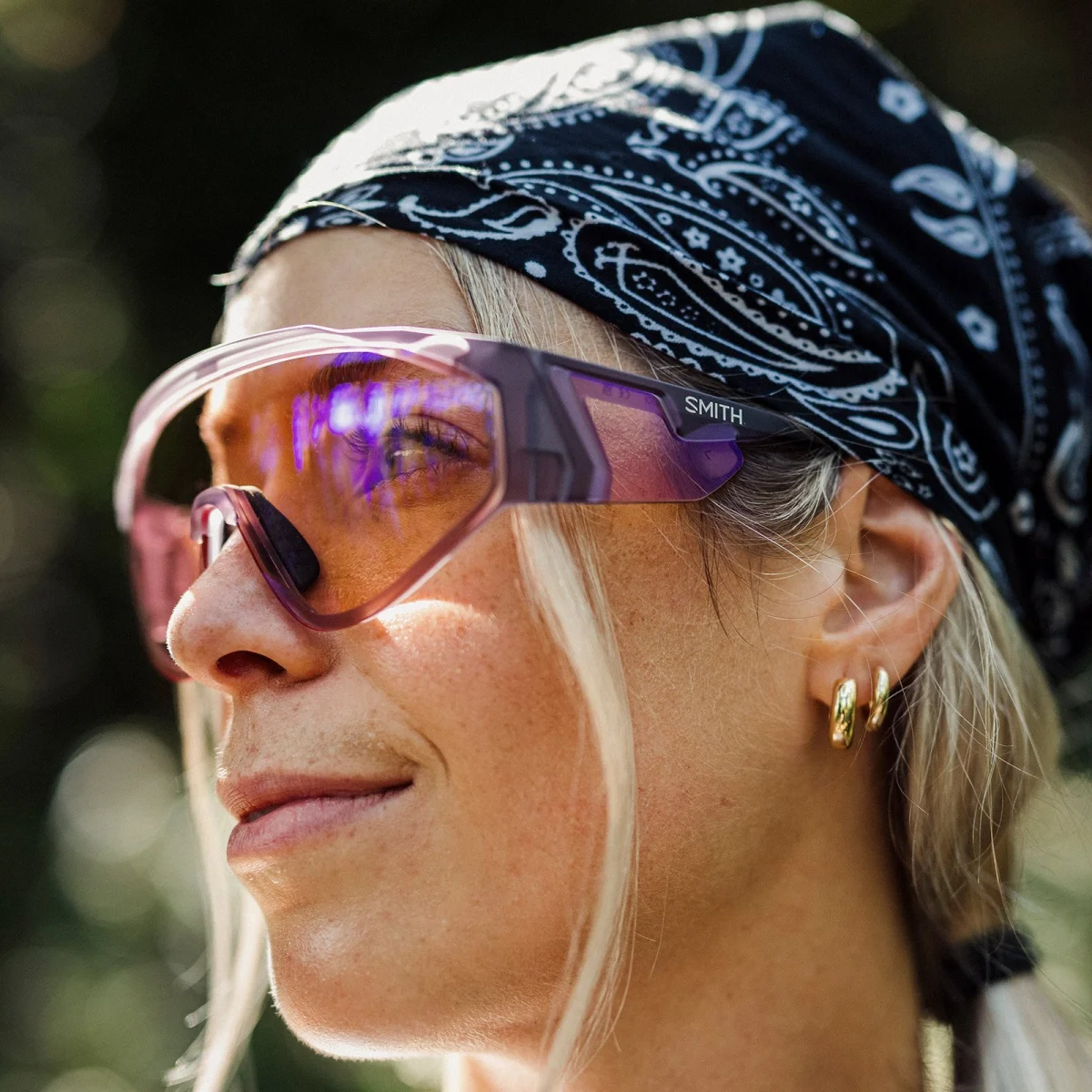 Person wearing large purple-tinted sunglasses, gold hoop earrings, and a black-and-white patterned bandana stands outdoors looking away.