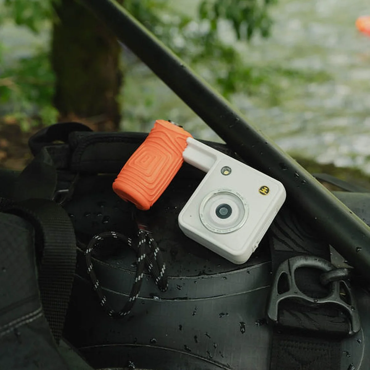 Compact camera with orange grip sitting on a wet black surface outdoors; water and greenery visible in the background.