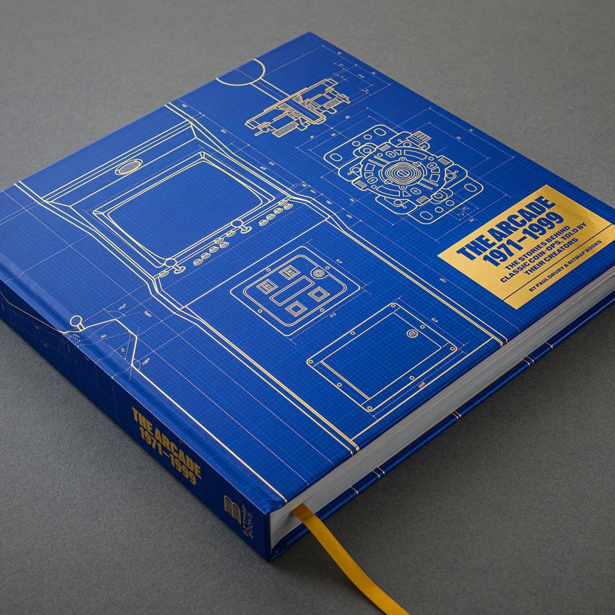 Hardcover book with blue cover, yellow technical drawings of arcade machines and controls, yellow title “The Arcade 1971–1996” on the front, and a visible yellow ribbon bookmark.