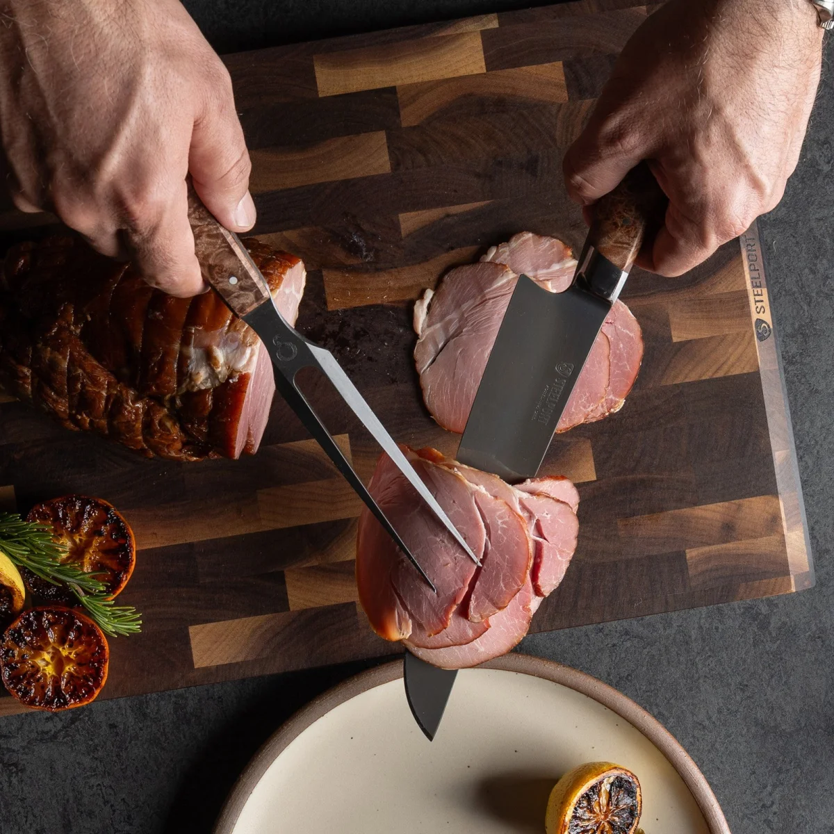 Sliced ham on a wooden cutting board is being cut with a knife and fork. Grilled lemon halves and fresh herbs are arranged beside the ham, with part of a plate visible at the bottom.