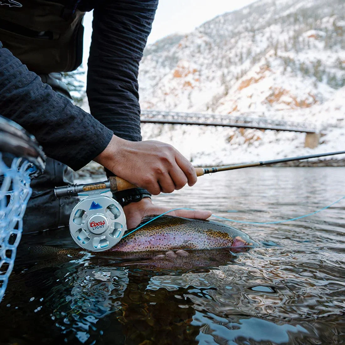 Person holding a fishing rod and a rainbow trout partially submerged in water with a snowy mountain and bridge in the background.