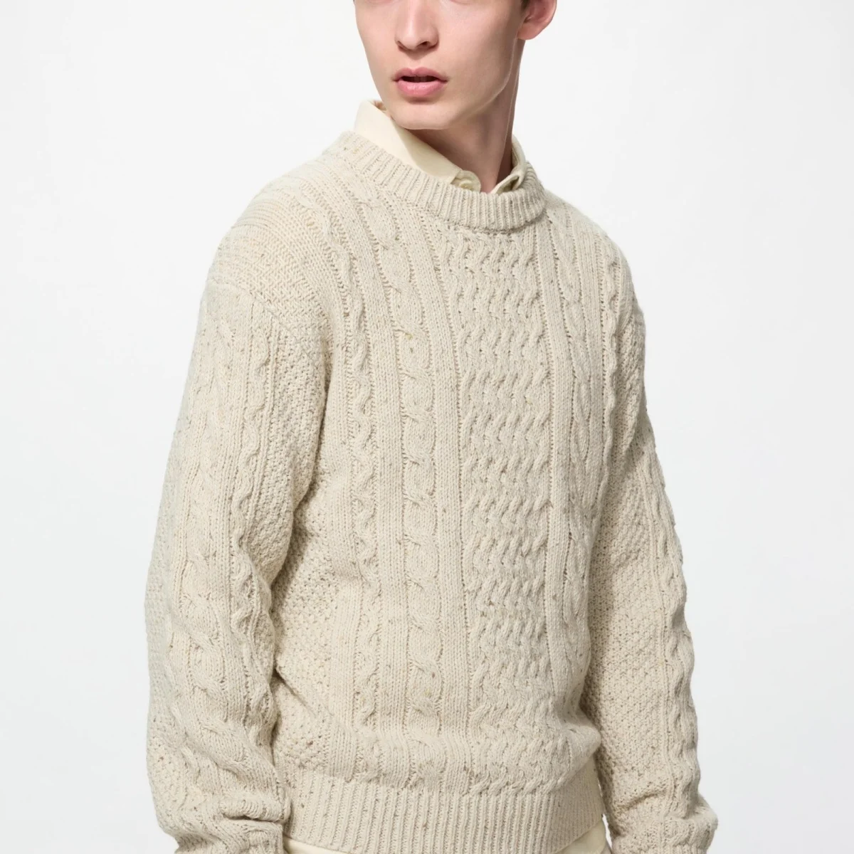 Cream-colored cable-knit sweater layered over a collared shirt, shown against a plain white background; person is standing and looking to the side.
