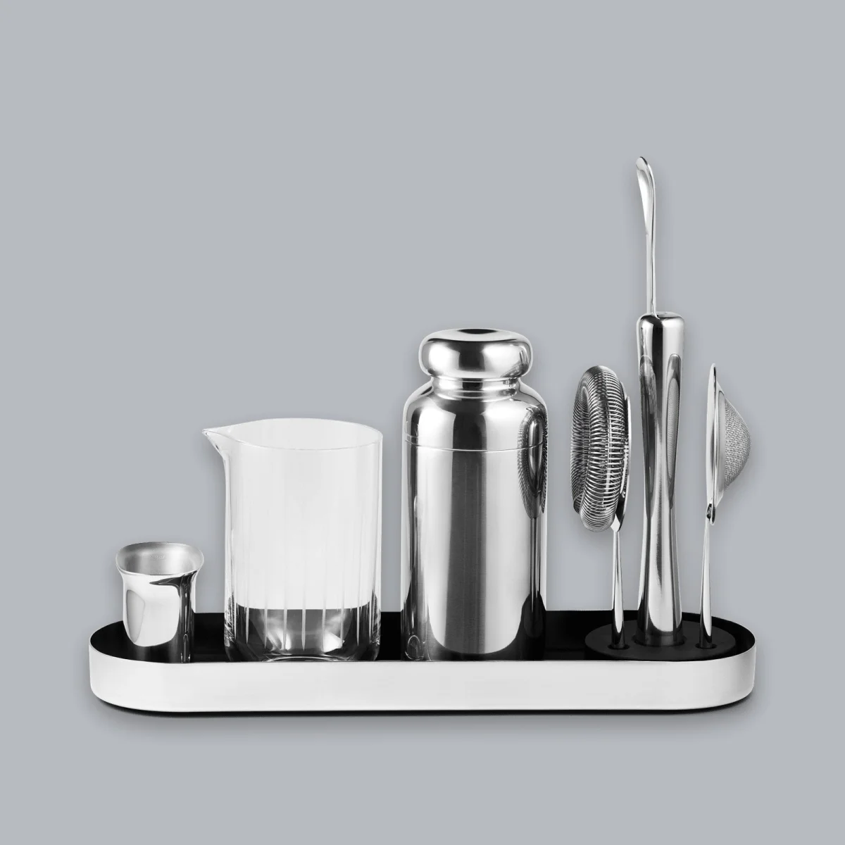 Stainless steel bar tool set on an oval black tray, including a jigger, glass mixing pitcher, shaker, strainer, bar spoon, and fine mesh sieve.