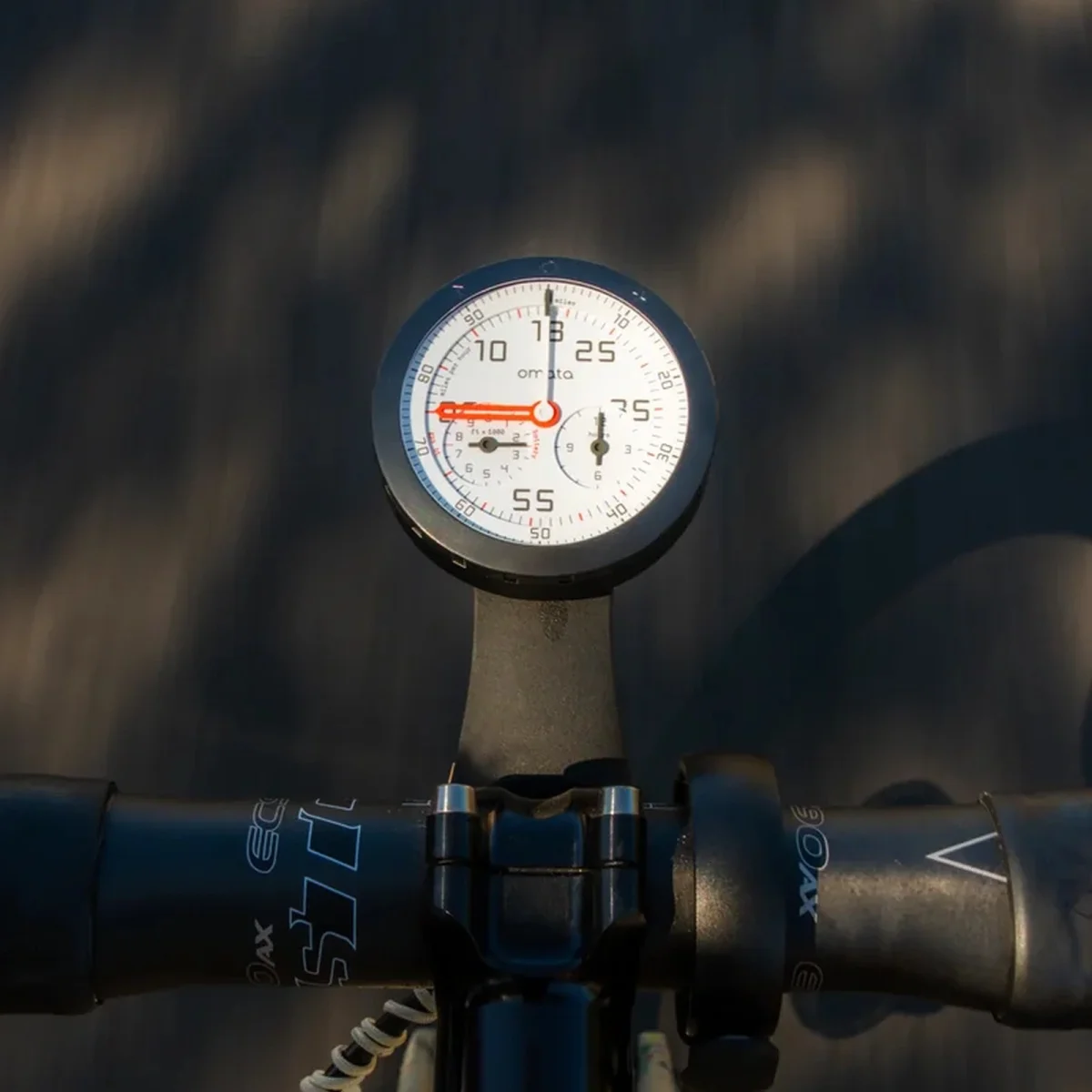 Omata analog cycling computer mounted on bicycle handlebars, set against a blurred road background.