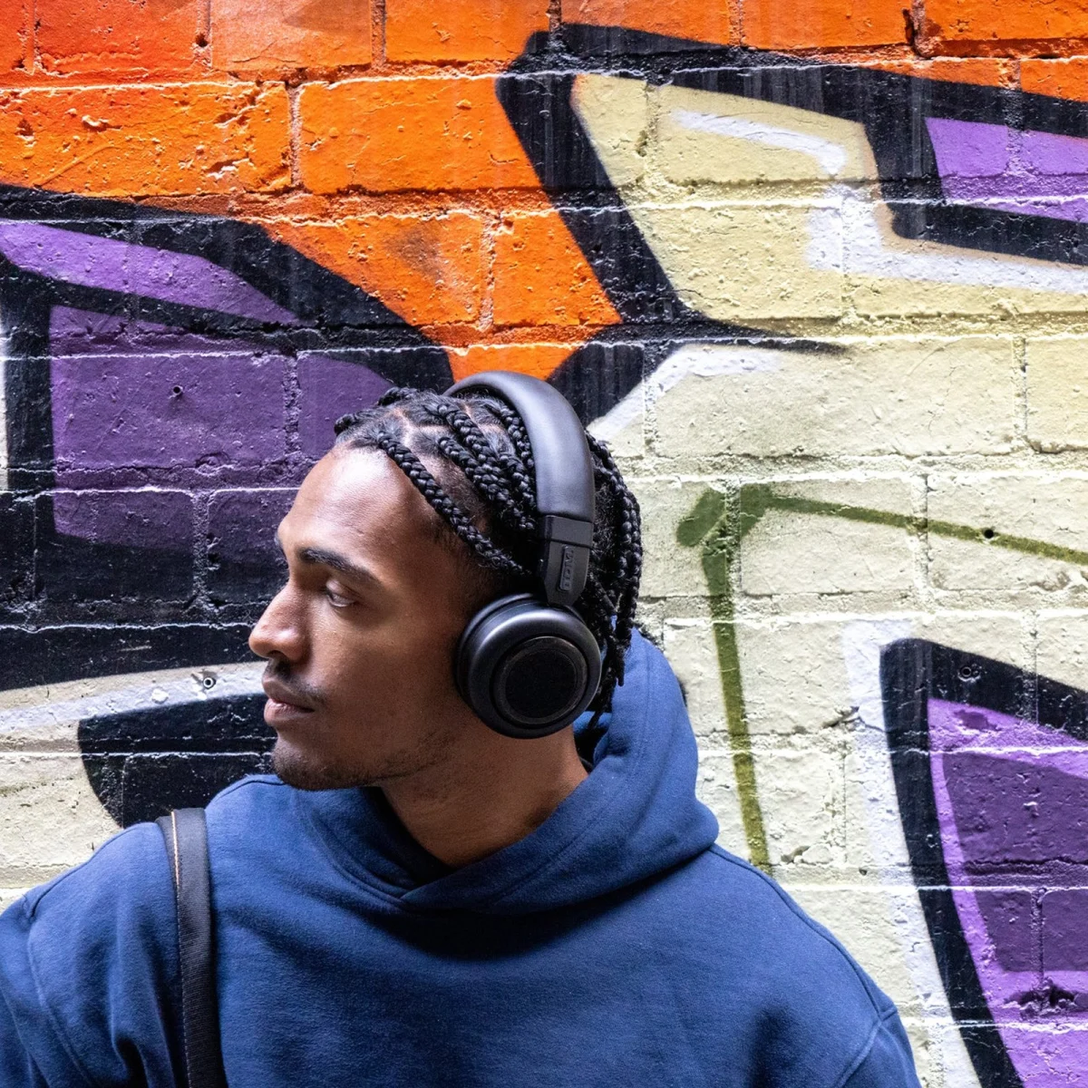 Man wearing black over-ear headphones and a blue hoodie in front of a colorful graffiti wall.