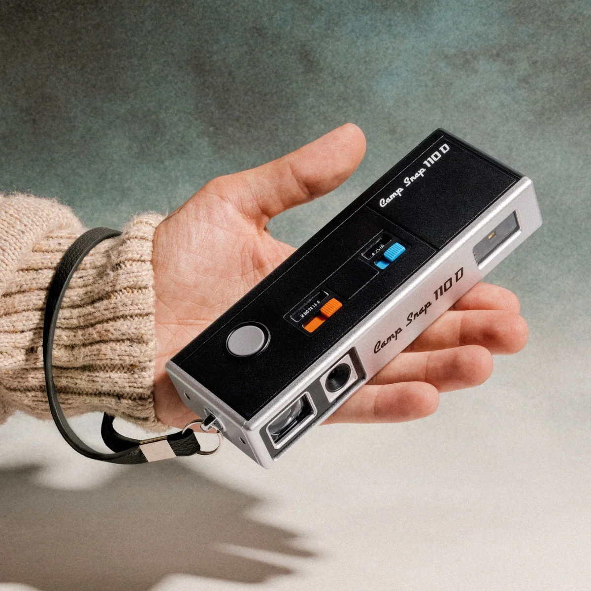 Vintage black and silver Carpo Snap 110D camera with colorful buttons and a wrist strap, held in a hand wearing a beige sweater, set against a green gradient background.