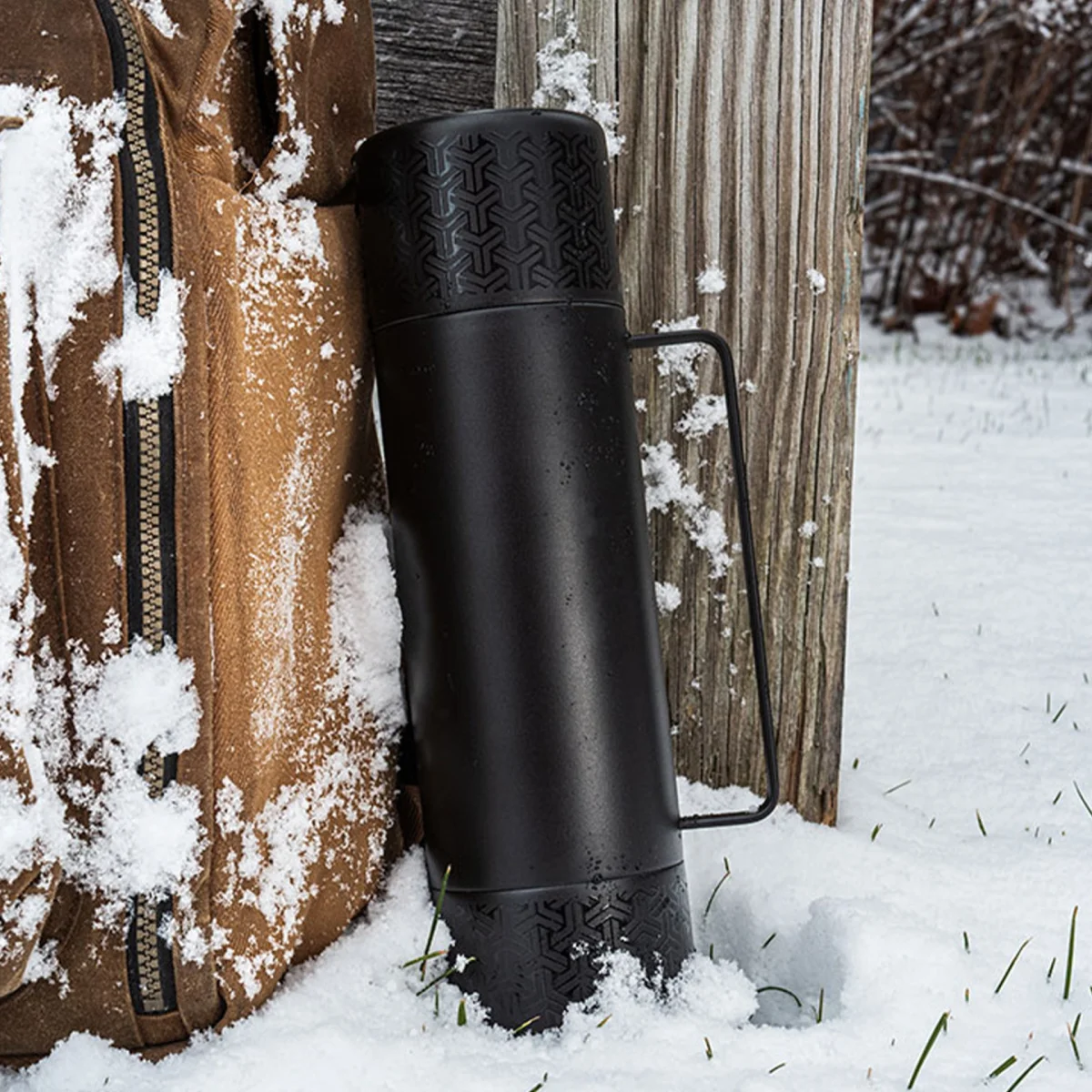 Black metal thermos with textured top and bottom and a handle, standing in snow next to a brown jacket and wooden post.