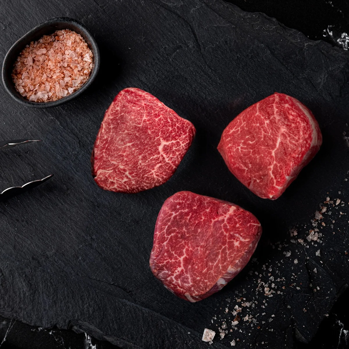 Three raw beef steaks arranged on a black stone slab, accompanied by a small bowl of pink salt and metal tongs.