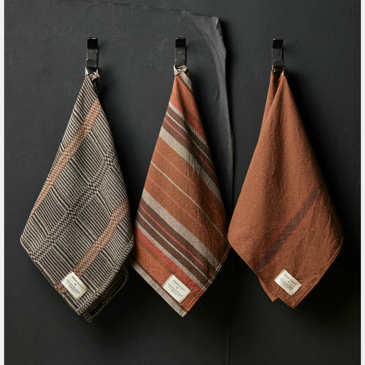 Three brown and beige patterned kitchen towels hanging on black metal hooks against a dark wall.