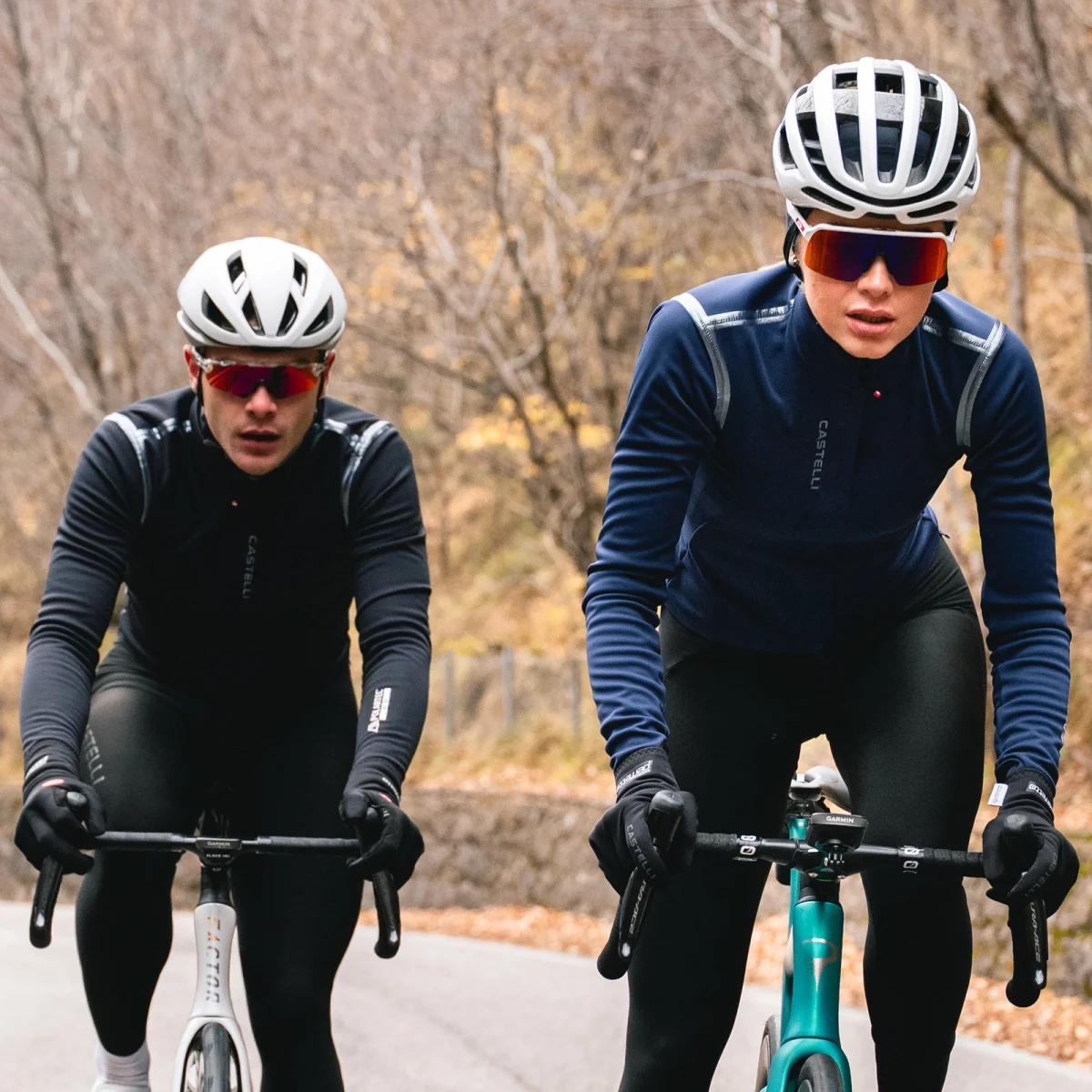 Two cyclists in helmets, sunglasses, and blue or black jackets ride along a road bordered by leafless trees in a cool outdoor setting.
