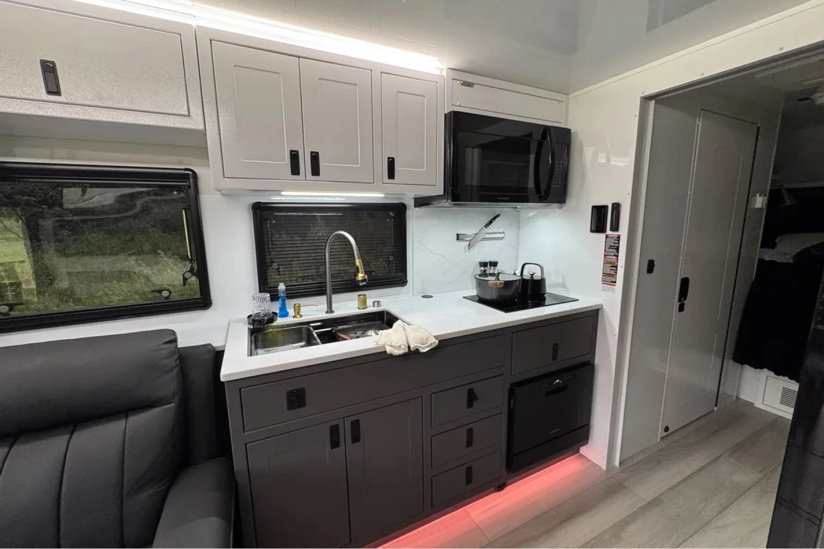 Camper kitchen interior with grey lower cabinets, light grey upper cabinets, a sink with a tall faucet, a microwave, and a black stovetop.