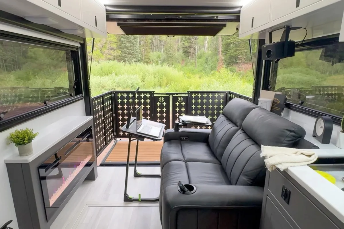 Black leather sofa, portable laptop desk, and electric fireplace inside a camper, positioned to face a rear patio deck with a forest view.