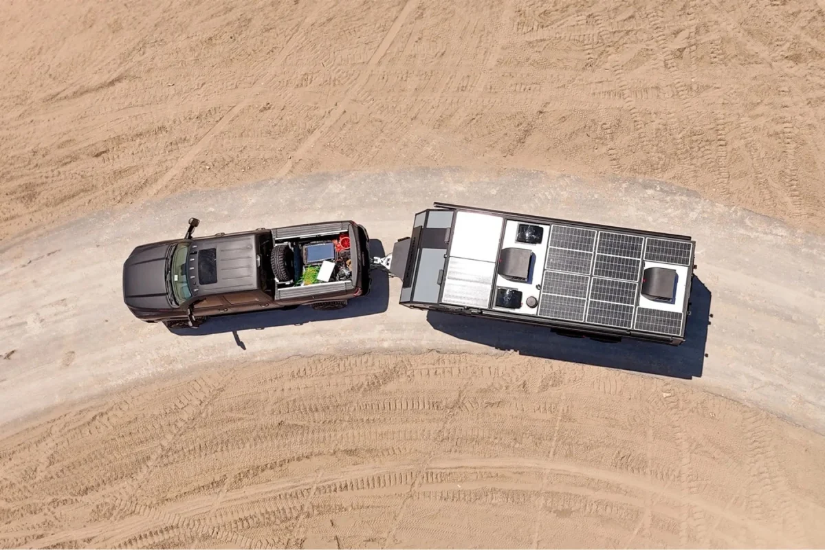 Aerial view of a dark pickup truck towing a black camper trailer with solar panels on its roof along a dirt path.