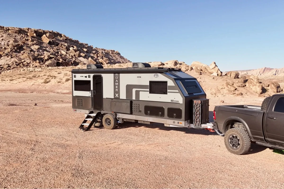 Grey and black Roamer X off-road travel trailer hitched to a black pickup truck, parked in a rocky desert landscape.