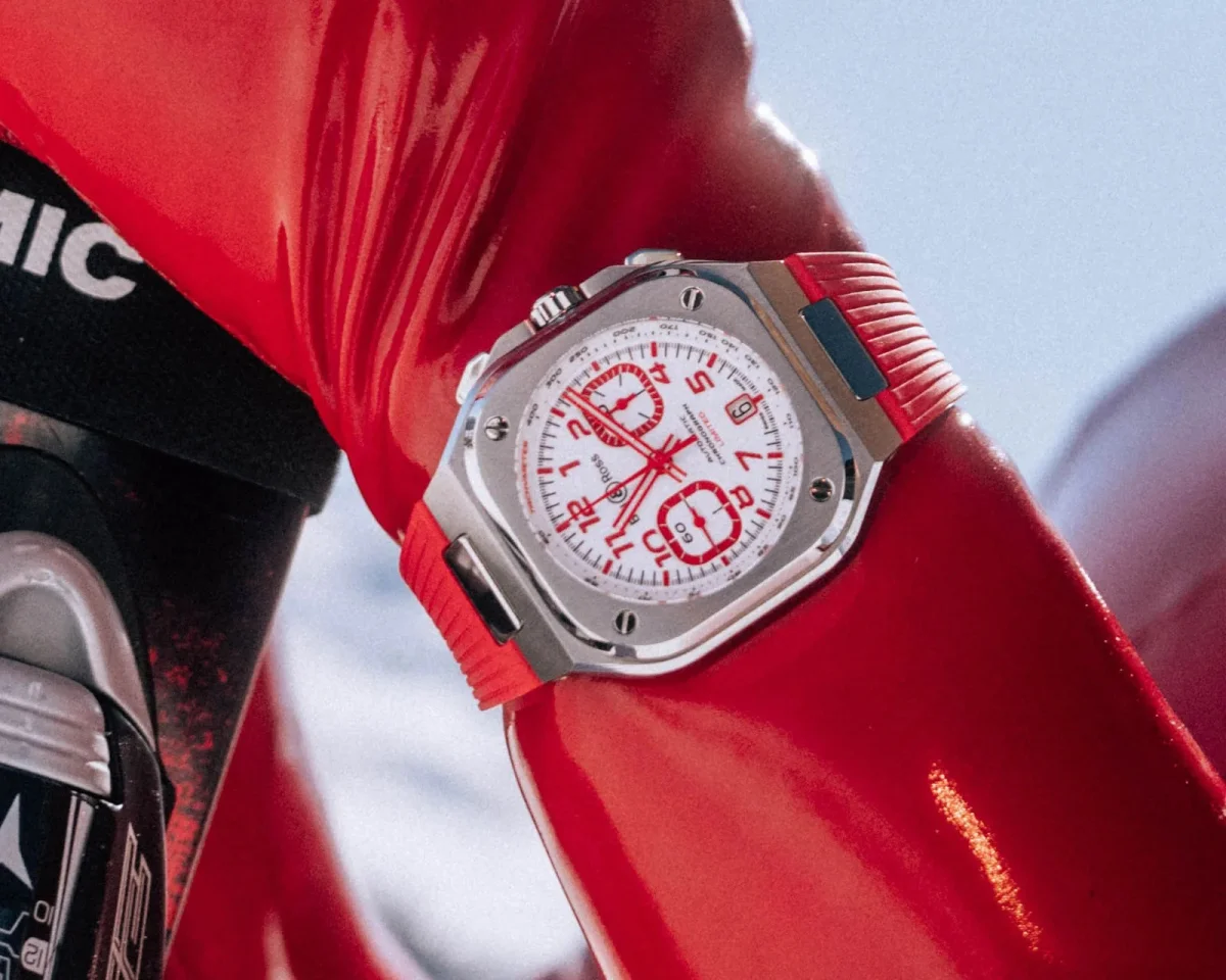 Bell & Ross chronograph watch featuring a square silver case, white dial, and red numerals, presented on a red rubber strap.