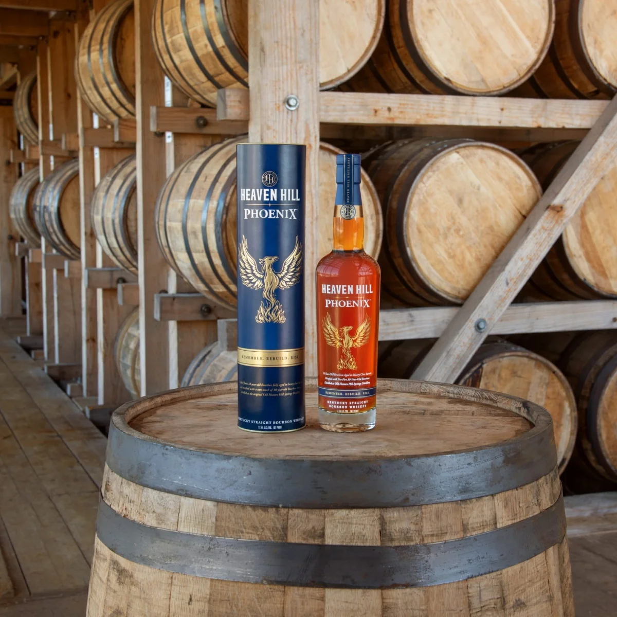Heaven Hill Phoenix Kentucky Straight Bourbon Whiskey bottle and its matching blue canister standing on a wooden barrel in a warehouse filled with stacked bourbon barrels.