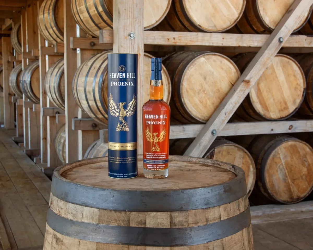 Heaven Hill Phoenix Kentucky Straight Bourbon Whiskey bottle and its matching blue cylindrical tube, standing on a wooden barrel in front of a rack of aging barrels.