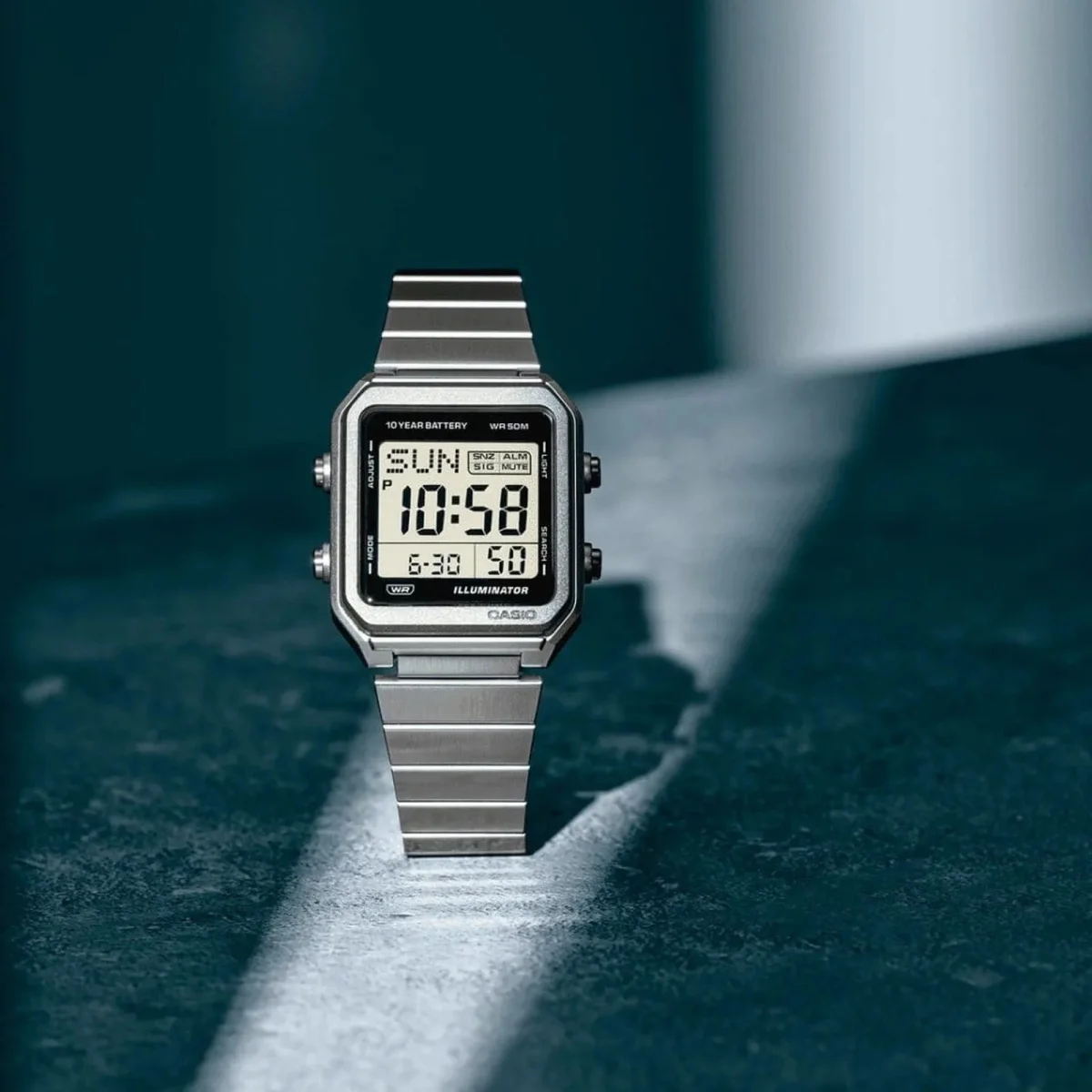 Silver Casio digital watch featuring a square face and a metal link band, displaying the time and date on a dark surface.