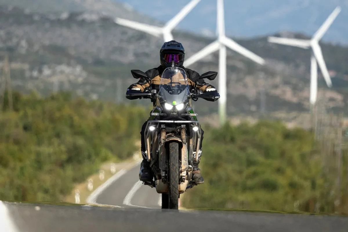 Dual-sport motorcycle being ridden on a paved road with wind turbines in the background.