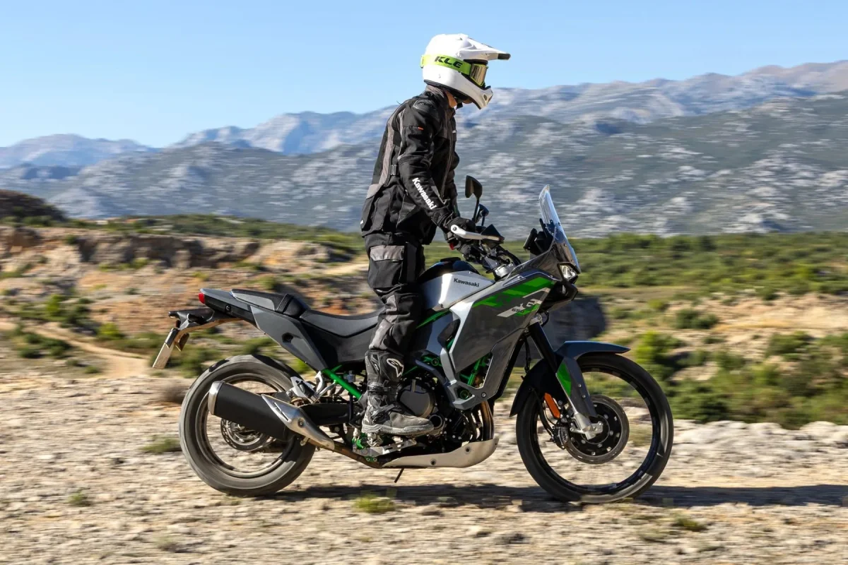 Grey and green Kawasaki Versys motorcycle with a rider in gear and helmet, traveling on a dirt trail with mountains in the background.