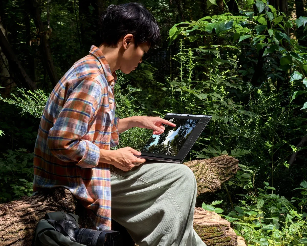 A plaid shirt worn while sitting on a log in a forest, with a hand touching the touchscreen of a laptop.