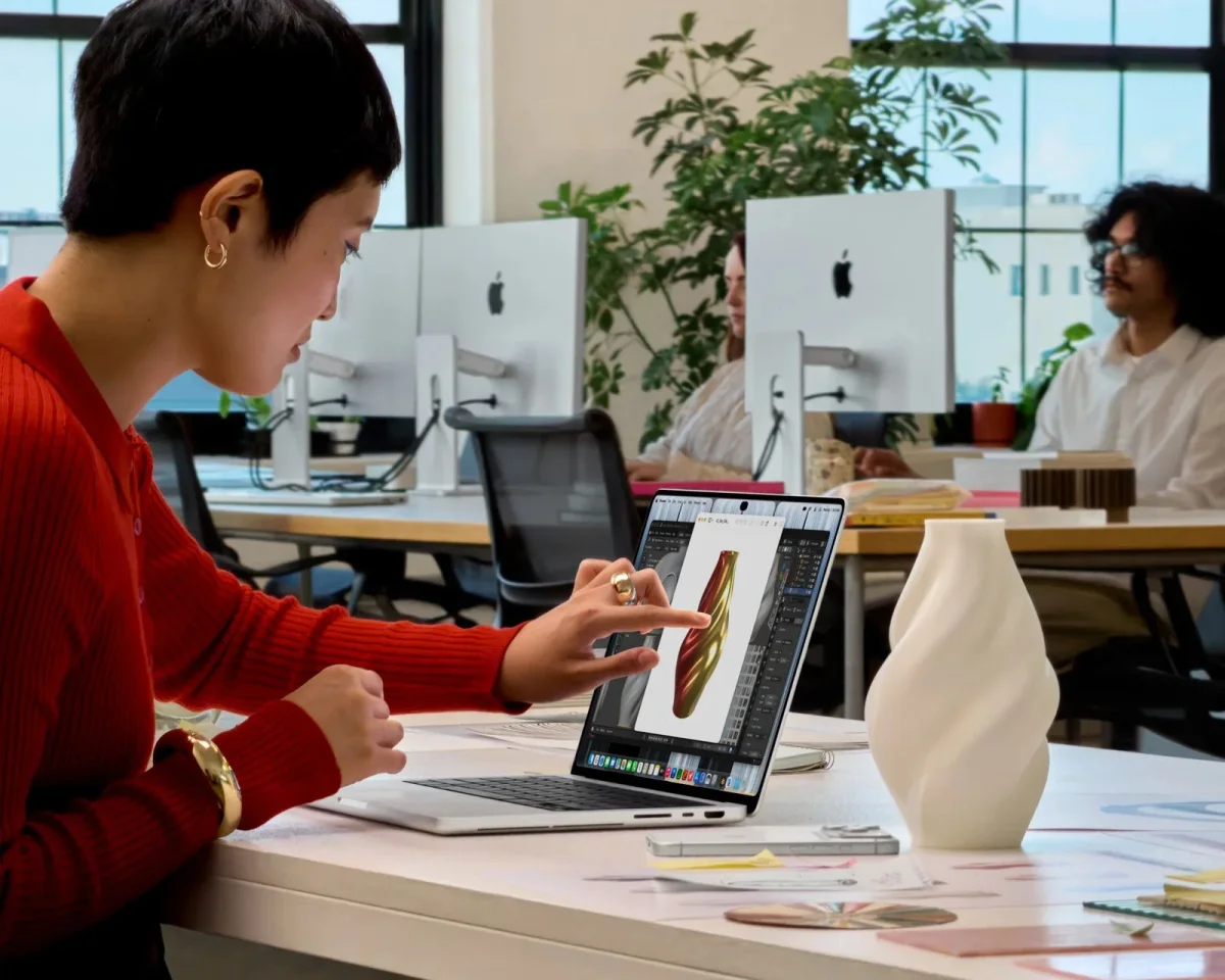 A finger interacts with a laptop screen displaying a 3D design in an office setting.