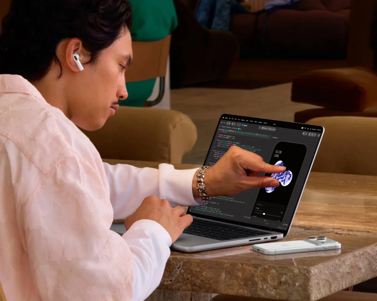 Wireless earbuds are worn while a finger interacts with a 3D graphic on a laptop screen, with a smartphone lying on the table nearby.