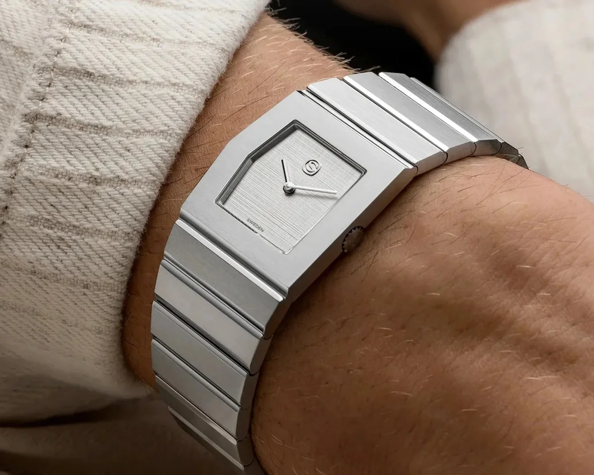 Silver-toned rectangular watch with an integrated link bracelet worn on a wrist.
