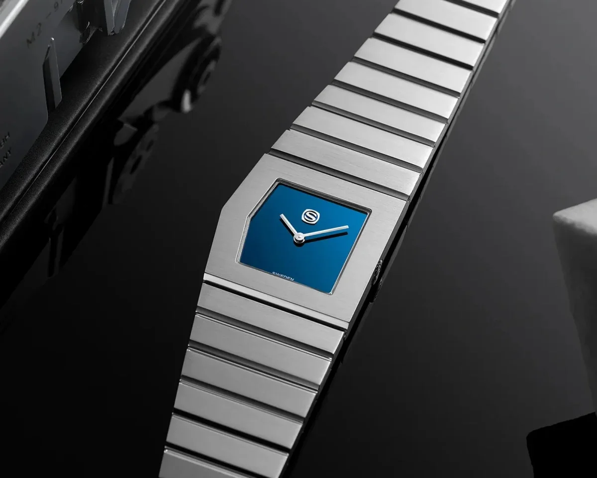 Silver-tone wristwatch with a square blue dial, white hands, and a tapered integrated metal link bracelet.
