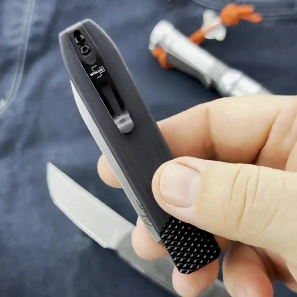 A closed black Boker pocket knife with a textured tail end and a pocket clip held in a hand, with an open knife and a small flashlight visible in the background.