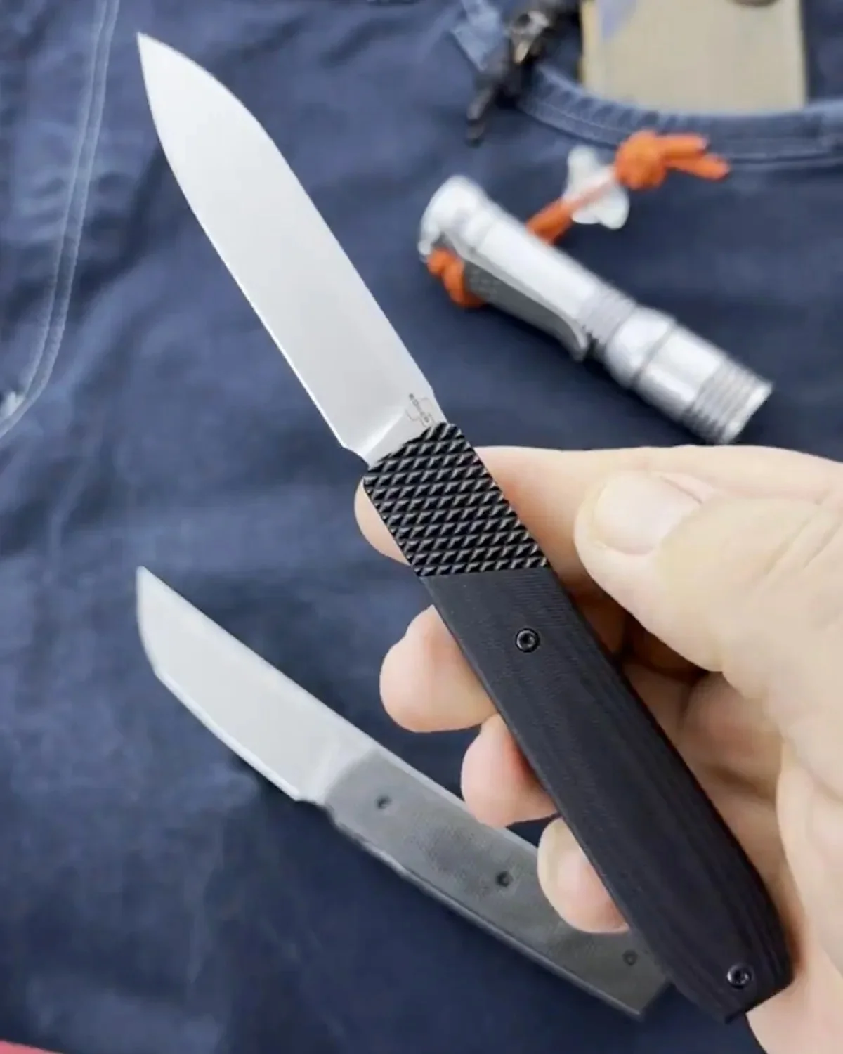 An open folding knife with a plain edge blade and a black textured handle held in a hand, with a second matching knife and a silver flashlight resting on blue fabric nearby.