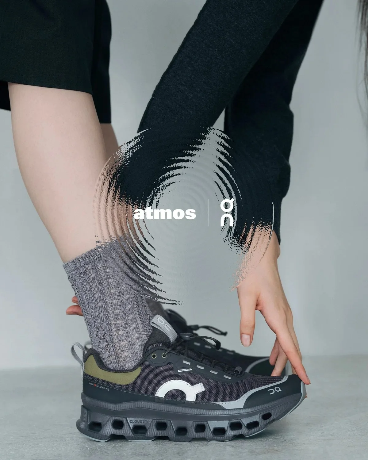 Black On Cloudultra sneaker with olive green accents and a patterned grey sock, held in a hand, featuring an overlaid atmos and On brand logo graphic.