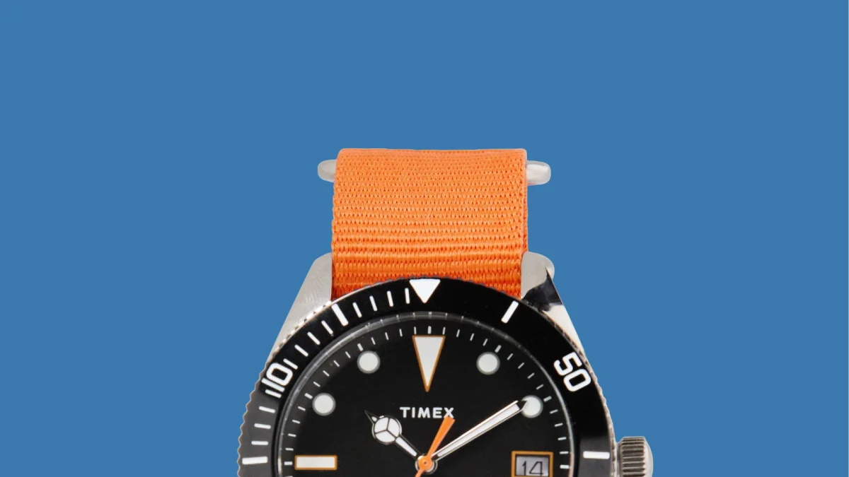 Timex watch with black dial, white markings, orange fabric strap, and visible date window showing 14, set against a solid blue background.