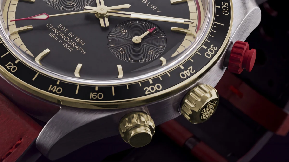 Close-up of a black chronograph watch face with gold markers, gold and red hands, gold crown and pushers, and a red crown on a red leather strap.