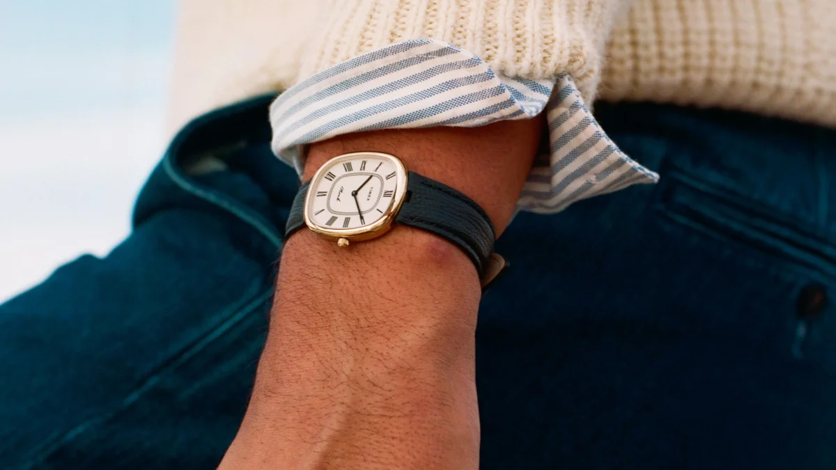Gold-tone cushion-shaped Timex watch with a white dial and black Roman numerals on a dark leather strap on a wrist.