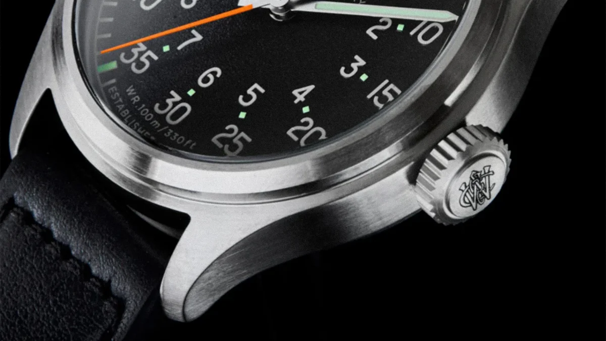 Close-up of a stainless steel watch case with a black dial, orange second hand, and black leather strap.