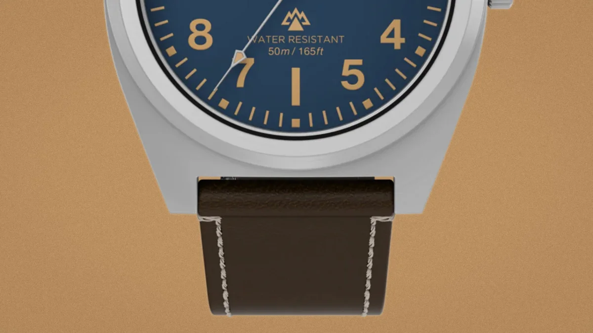 Close-up of a watch with a blue dial, gold numerals, silver case, and dark brown leather strap with white stitching.