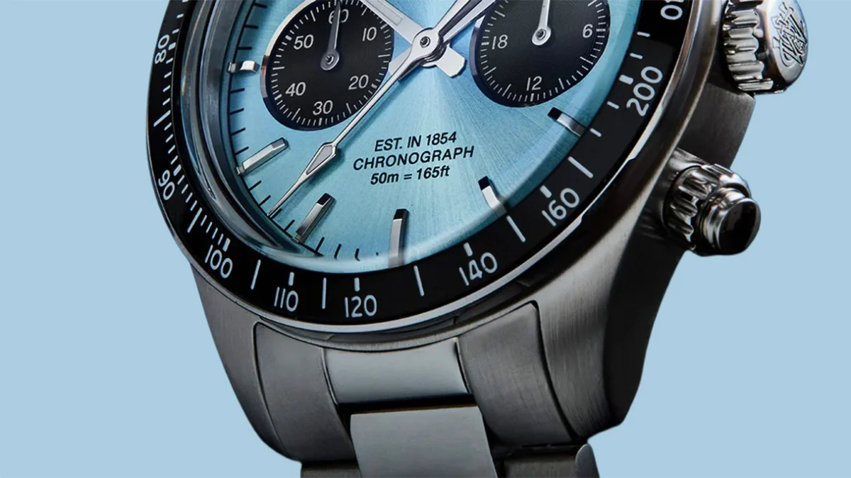 Close-up of a stainless steel chronograph watch with a light blue dial and black subdials on a light blue background.