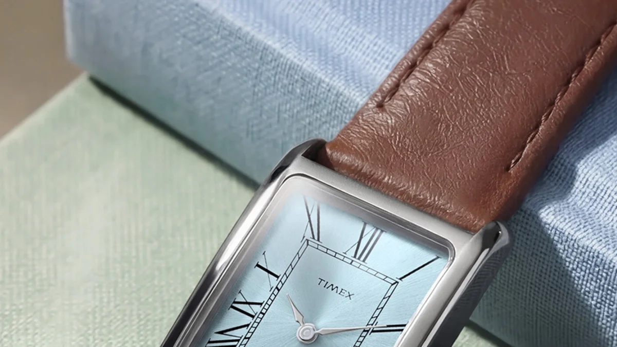 Rectangular silver Timex watch with light blue dial and brown leather strap on textured fabric.