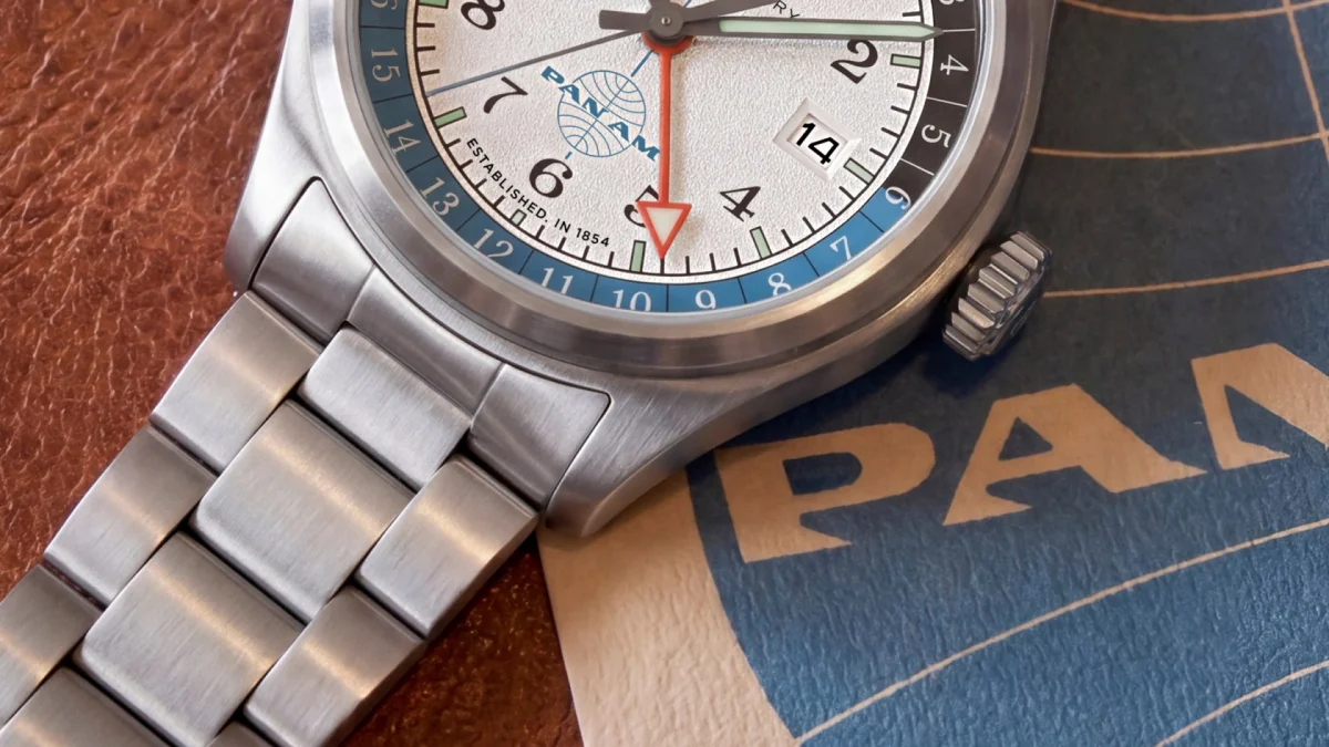 Pan Am branded wristwatch with a white dial, stainless steel bracelet, and GMT hand, resting on a textured surface.