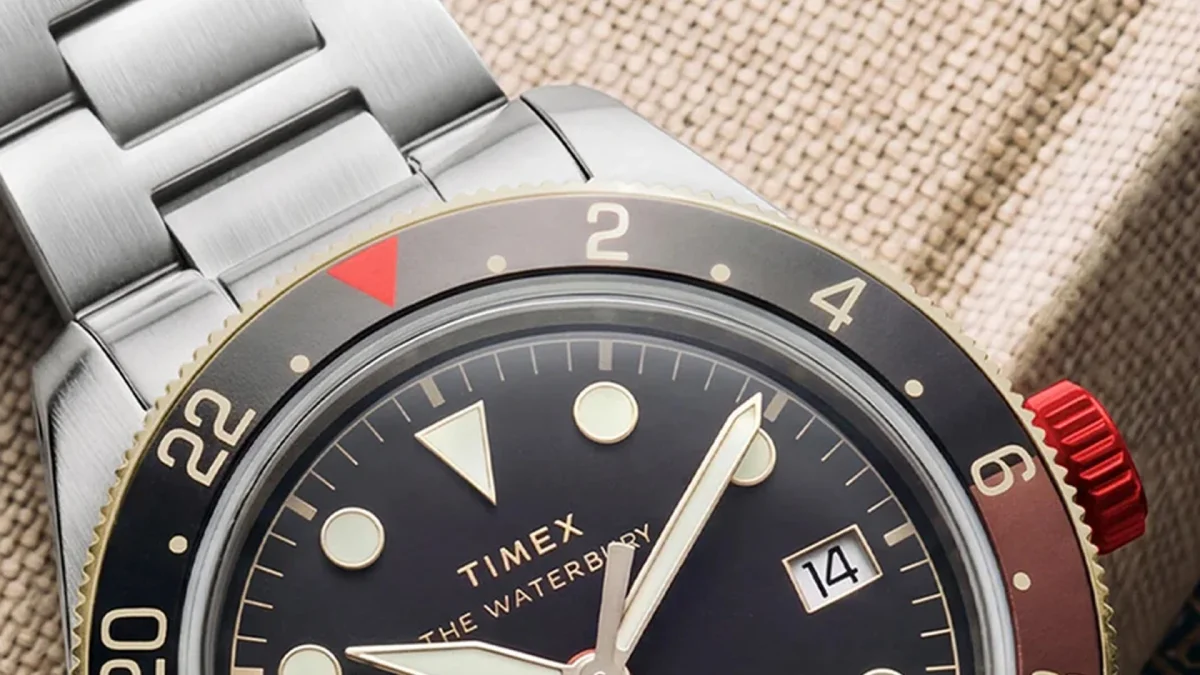 Timex wristwatch with a black face, bold hour markers, date window at 3 o’clock, two-tone bezel, and stainless steel band on a textured fabric surface.